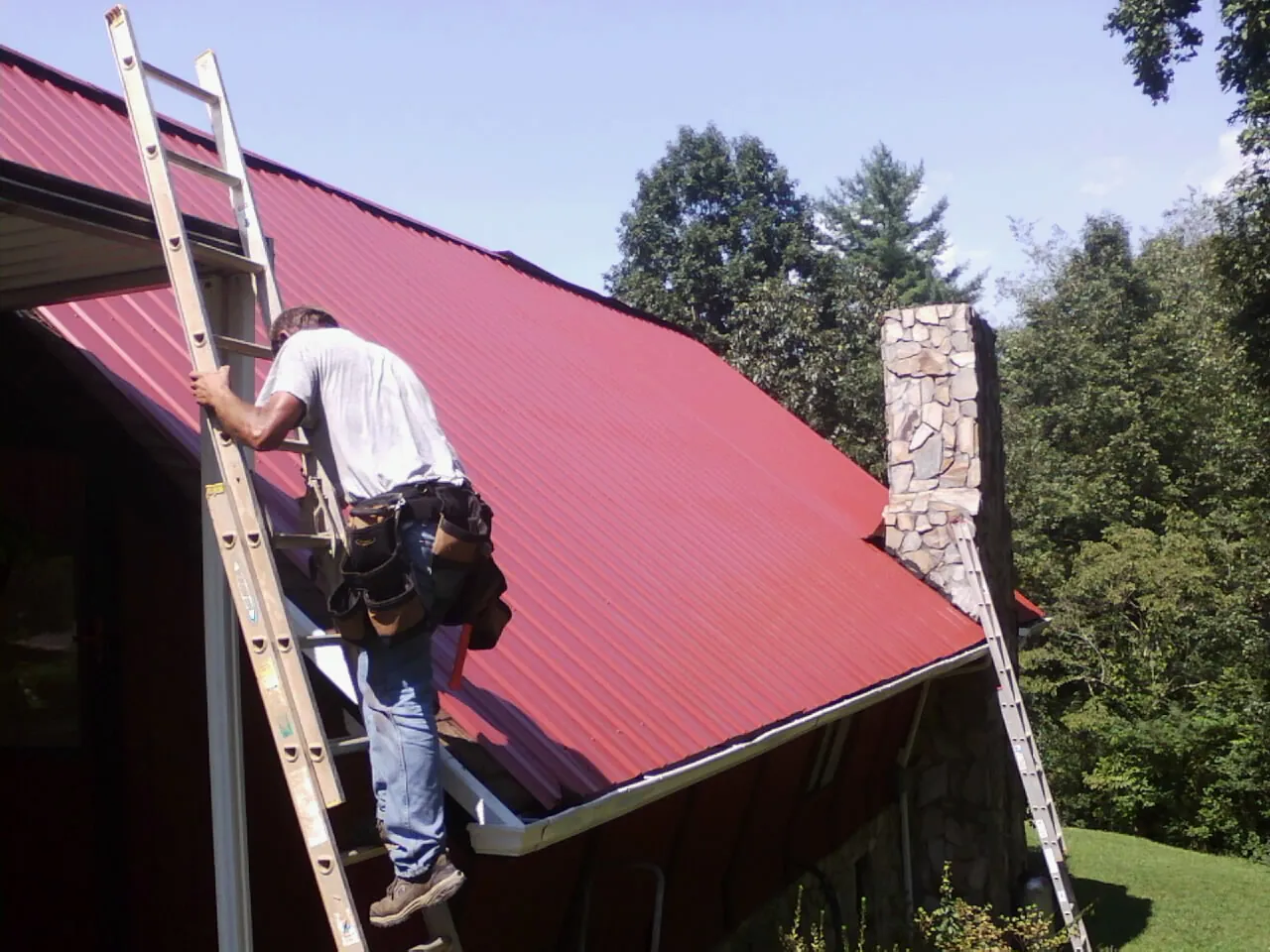 Roofer on a ladder beside a red metal roof and stone chimney — Soffit & Fascia Repair by Roofing Brothers in Damascus